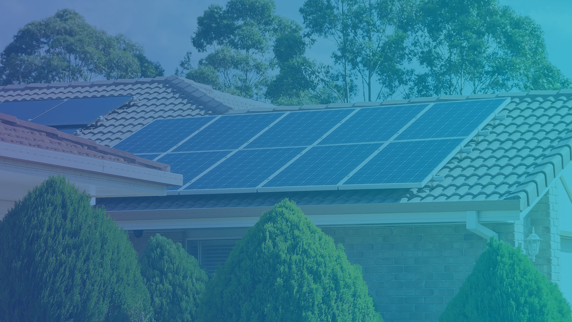 Das Bild zeigt die Dächer eines Hauses, auf denen mehrere Photovoltaikmodule installiert sind. Die Module sind in Reihen angeordnet und decken einen großen Teil der Dachfläche ab. Die Dächer bestehen aus klassischen Ziegeln, und die Solaranlage ist so positioniert, dass sie eine optimale Sonneneinstrahlung erhält. Im Vordergrund sind mehrere grüne, gepflegte Büsche zu sehen, die das Haus umgeben. Im Hintergrund ragen hohe Bäume empor, die eine natürliche Kulisse bilden. Die Szene vermittelt eine nachhaltige und umweltbewusste Wohnumgebung, in der erneuerbare Energien eine zentrale Rolle spielen. Die Solaranlage könnte in ein Smart-Home-System eingebunden sein, um den erzeugten Strom effizient zu verwalten und den Energieverbrauch im Haushalt zu optimieren.