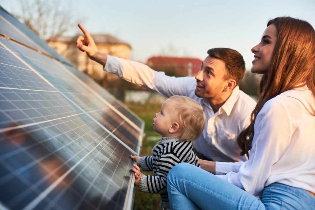 Ein Mann zeigt seiner Familie die Solarpaneele auf dem Grundstück in der Nähe des Hauses an einem warmen Tag. Eine junge Frau mit einem Kind und ein Mann stehen im Sonnenlicht und betrachten die Solarpaneele.