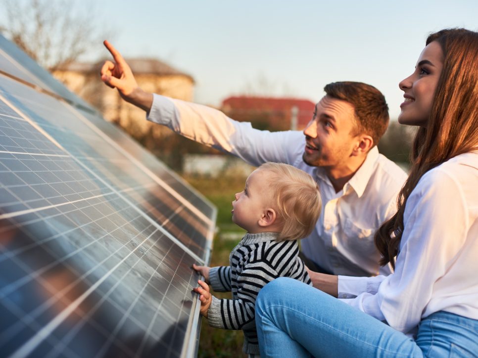 Ein Mann zeigt seiner Familie die Solarpaneele auf dem Grundstück in der Nähe des Hauses an einem warmen Tag. Eine junge Frau mit einem Kind und ein Mann stehen im Sonnenlicht und betrachten die Solarpaneele.