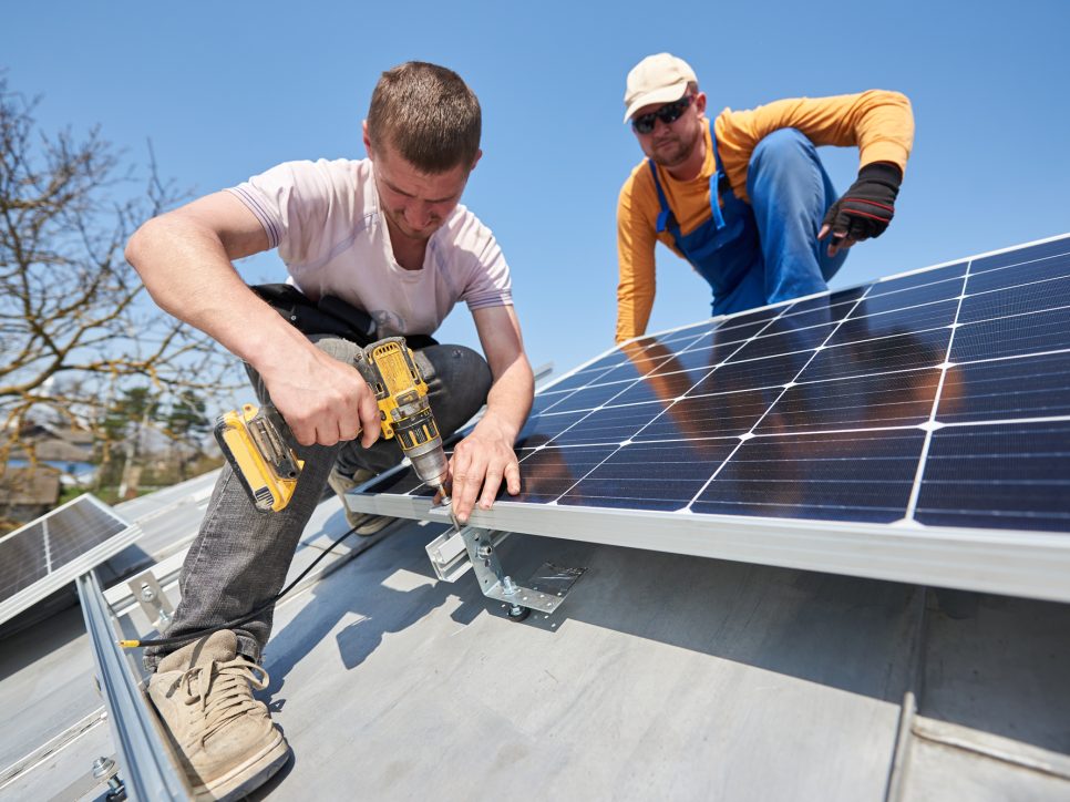 Male engineers installing solar photovoltaic panel system using screwdriver. Two electricians mounting solar module on roof of modern house. Alternative renewable innovation concept.