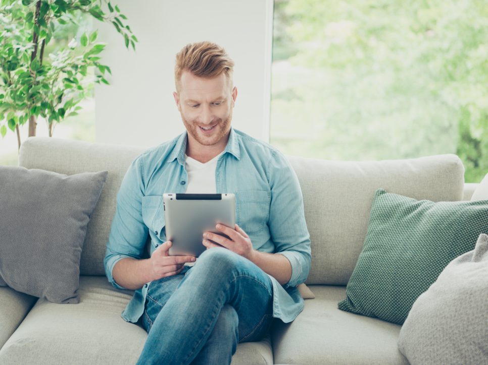 Attractive handsome good-wearing man in stylish trendy casual denim shirt jeans look at tablet screen enjoy movie or entertainment show sit on cozy sofa indoor bright light apartment
