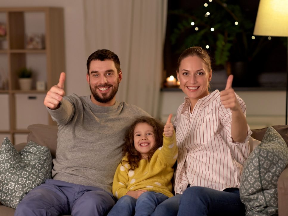 family and people concept - portrait of happy smiling father, mother and little daughter sitting on sofa and showing thumbs up at home at night