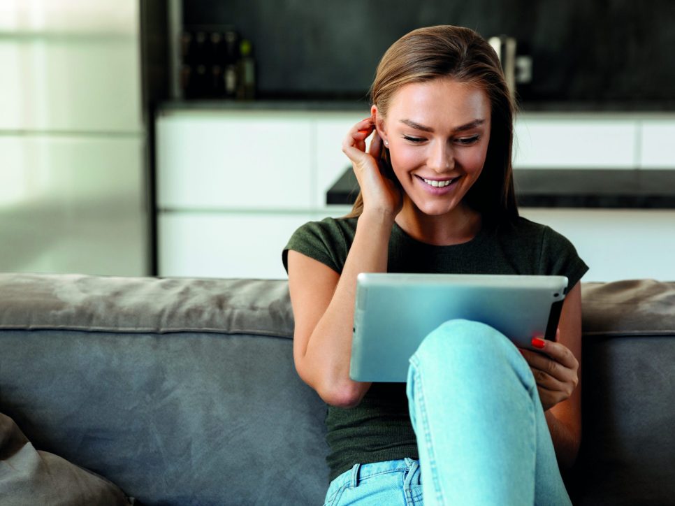 Smiling young woman sitting on a couch at home, using tablet computer