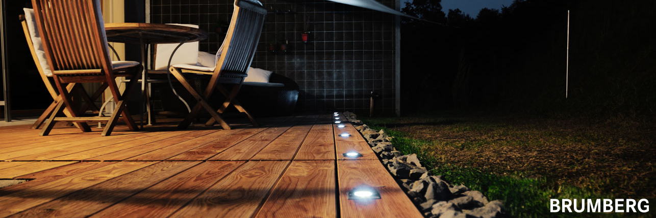 Stimmungsvolle Terrassenbeleuchtung mit Brumberg-Einbauleuchten – Eine gemütliche Holzterrasse mit integrierter LED-Bodenbeleuchtung von Brumberg. Die eingebauten Spots sorgen für eine dezente, blendfreie Ausleuchtung entlang der Terrassenkante. Ein Holz-Esstisch mit passenden Stühlen steht bereit für gesellige Abende im Freien. Im Hintergrund eine moderne Hausfassade mit bepflanzter Wand. Die warmweiße Beleuchtung schafft eine einladende Atmosphäre bei Nacht.