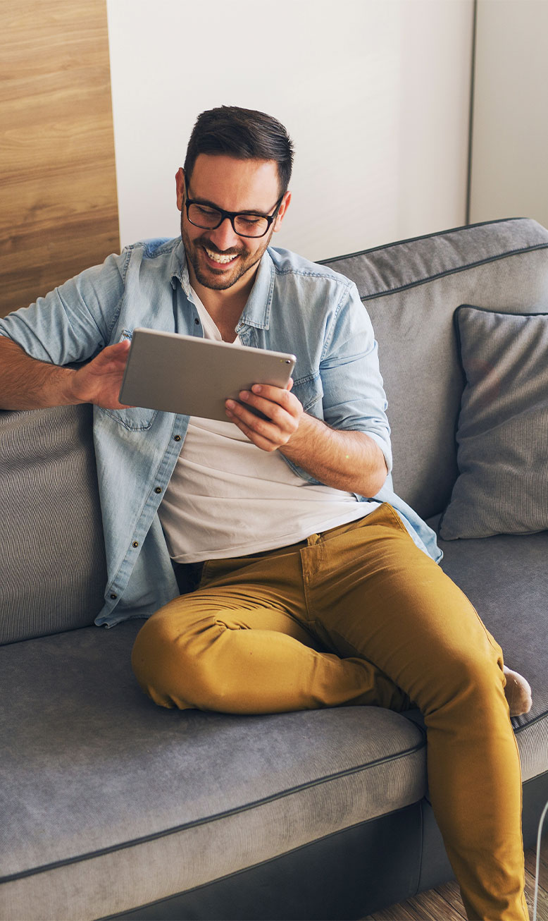 Lächelnder Mann mit dunklem Haar und Brille sitzt auf grauem Sofa, lässig gekleidet in hellblauem Jeanshemd über weißem T-Shirt und senffarbener Hose, steuert sein SmartHome entspannt über ein Tablet.