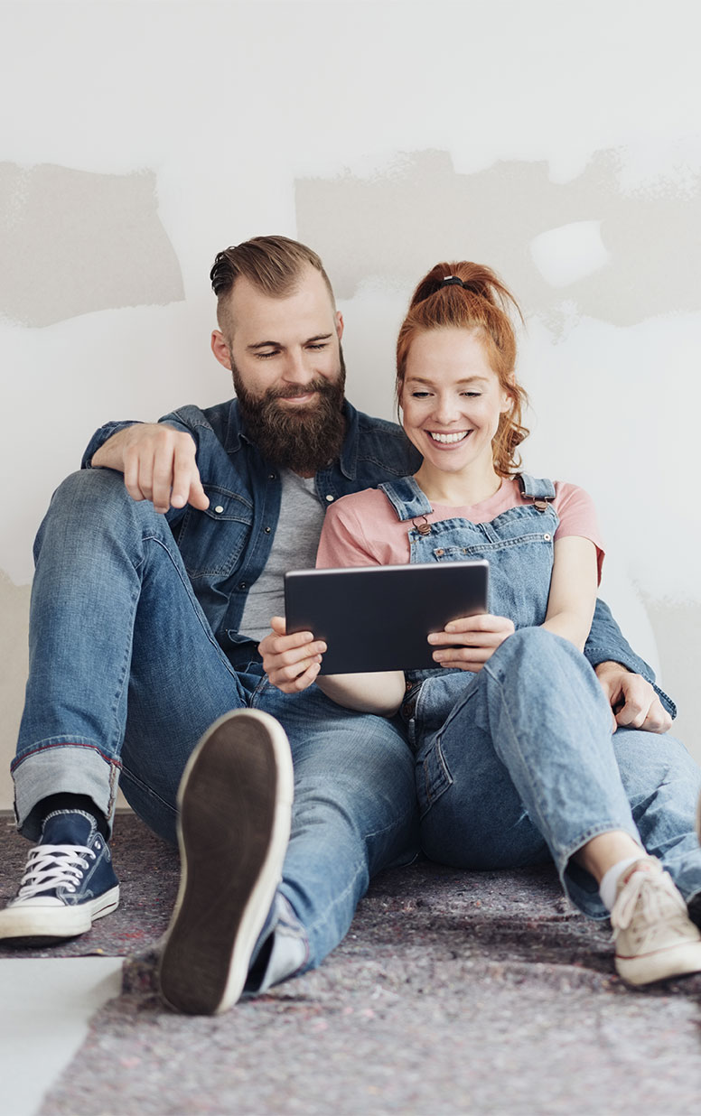 Mann mit Bart und Frau mit roten Haaren sitzen auf dem Boden vor strukturierter Wand, beide in Jeans-Outfits. Sie lächeln und schauen auf ein Tablet, das die Frau hält, während sie SmartHome-Möglichkeiten erkunden. Der Mann zeigt aufgeregt auf den Bildschirm, beide strecken bequem die Beine aus.