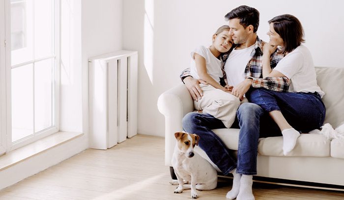 Dreiköpfige Familie sitzt mit ihrem Hund auf einem Sofa in einem hellen SmartHome-Wohnzimmer. Eltern umarmen liebevoll ihr kleines Kind, während der weiß braune Hund neben dem Sofa das gemütliche Ambiente genießt, das durch die Hausautomation der KNX-Systeme geschaffen wird.