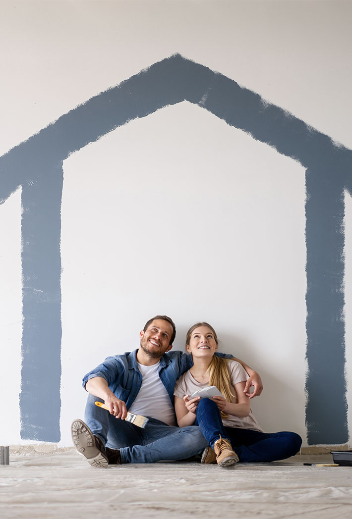 Paar sitzt auf dem Boden und lehnt an einer Wand mit blau gestrichenen Hausumrissen. Der Mann hält einen Pinsel. Der Raum wird renoviert oder neu gestrichen, zur Integration von SmartHome Funktionen für mehr Komfort.