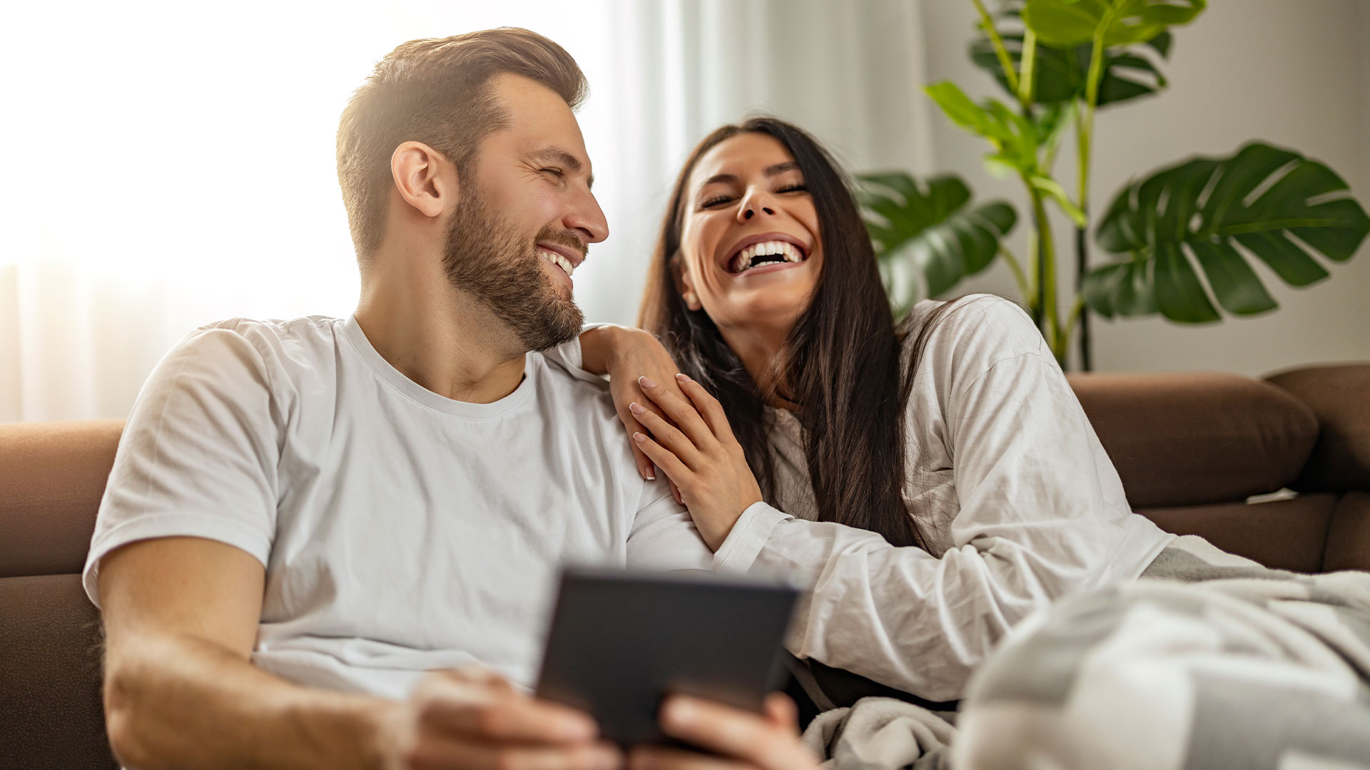 Ein lächelnder Mann und eine lächelnde Frau sitzen zusammen auf einem Sofa und lachen. Der Mann hält ein Tablet, auf dem ihre SmartHome-Einstellungen angezeigt werden, während die Frau an seiner Schulter lehnt. Sie sind leger gekleidet, im Hintergrund sind eine Zimmerpflanze und Sonnenlicht zu sehen.