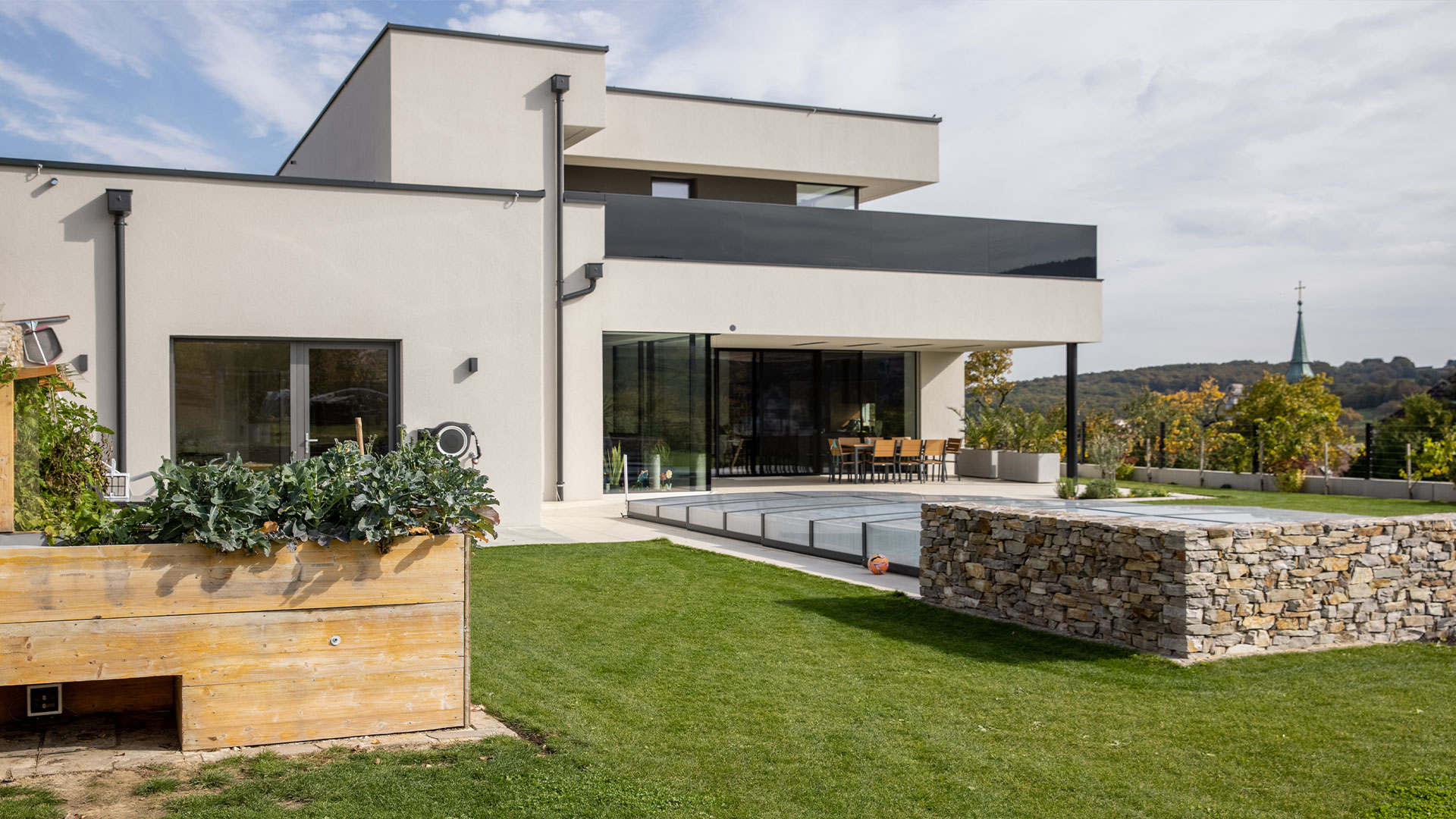 Modernes zweistöckiges Haus mit minimalistischem Design, Flachdächern und großen Fenstern, ergänzt durch eine geräumige Terrasse. Im Vordergrund befindet sich ein erhöhtes Gartenbeet aus Holz mit Pflanzen und einer Steinmauer. Die Terrasse verfügt über einen Essbereich, einen eingebauten Pool und fortschrittliche Hausautomation durch KNX-Systeme.