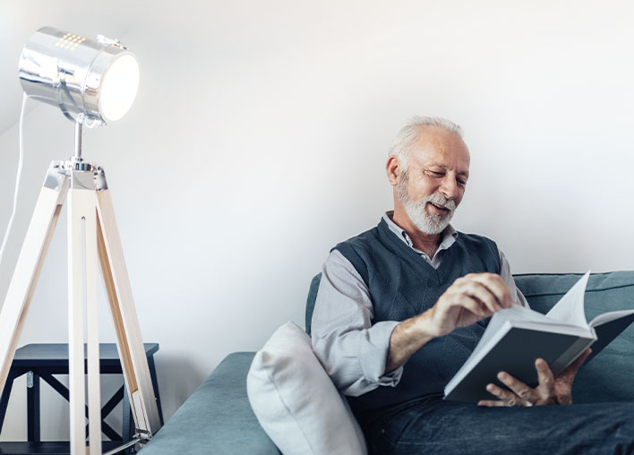 Älterer Mann sitzt entspannt auf einem Sofa und liest ein Buch, während eine stilvolle Stehlampe für angenehme Beleuchtung sorgt. Das SmartHome-System "TONI" passt die Beleuchtung automatisch an, um optimalen Komfort und Lesebedingungen zu gewährleisten.