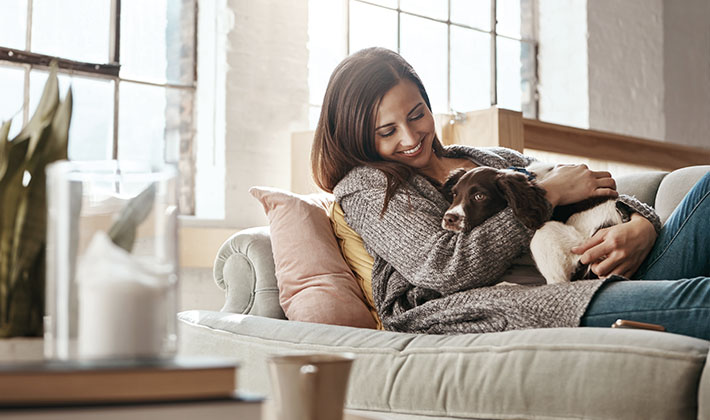 Frau sitzt entspannt auf einem Sofa in einem gemütlichen Wohnzimmer, hält einen Hund im Arm und lächelt glücklich. Das Smart Home sorgt für eine behagliche Atmosphäre durch smarte Beleuchtung und Klimasteuerung.