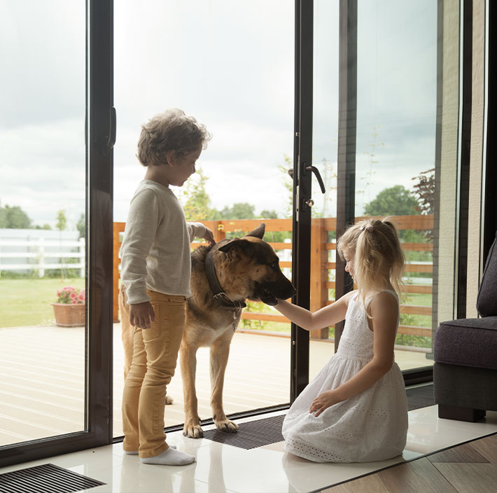 Zwei Kinder spielen mit einem Schäferhund vor einer großen Glasschiebetür, die zu einer Terrasse führt. Das helle, moderne Zuhause nutzt SmartHome-Technologie für komfortable Hausautomation, während die Kinder die sichere und gemütliche Umgebung genießen.