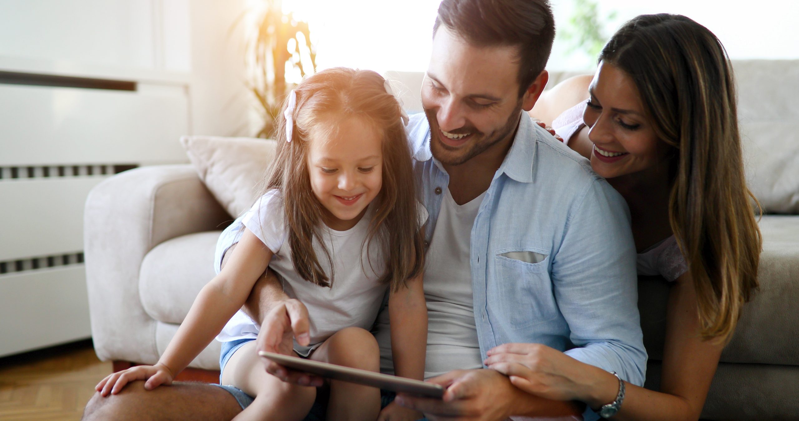 Junge Familie sitzt im Wohnzimmer auf dem Boden und schaut lächelnd auf ein Tablet. Vater im blauen Hemd präsentiert das Smart Home-Setup, während Mutter und Tochter eng zusammengekuschelt den Bildschirm betrachten