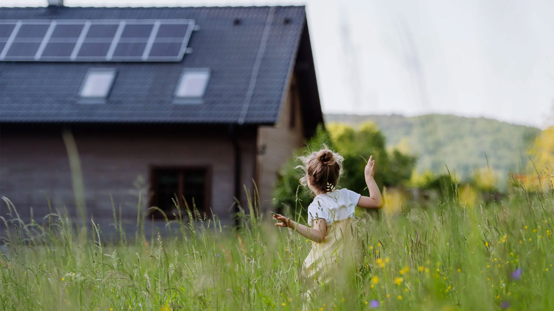 Junges Mädchen mit blondem Haarknoten läuft durch hohe Wiese, SmartHome mit KNX-Systemen und Solarpanelen im Hintergrund, umgeben von grünen Hügeln und Bäumen unter klarem Himmel.