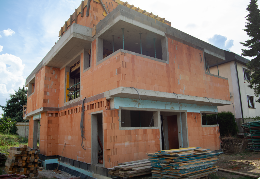 Das Bild zeigt den Rohbau eines zweistöckigen Hauses aus roten Ziegeln. Der Bau befindet sich noch in einer frühen Phase, da das Gebäude noch keine Fenster, Türen oder Außenverkleidung hat. Einige Baustellenmaterialien wie Holz und Gerüstteile liegen rund um das Haus verstreut. An der Außenwand sind einige Kabel sichtbar, was darauf hindeutet, dass elektrische Installationen vorbereitet werden. Es ist möglich, dass dieses Gebäude später als Smart Home ausgestattet wird, da solche Installationen oft schon im Rohbau geplant werden.