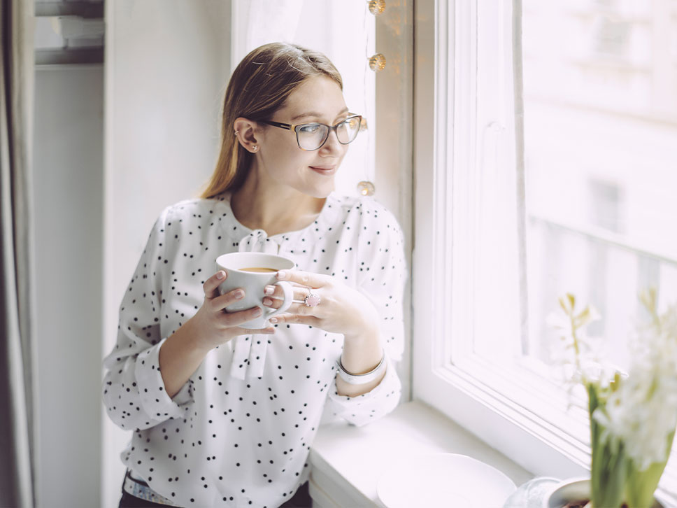 Person mit Brille und gepunkteter weißer Bluse steht am Fenster, hält eine Tasse Kaffee oder Tee und lächelt sanft nach draußen. Genießt die helle, luftige Atmosphäre, die durch das SmartHome-System geschaffen wird. Licht fällt durch das Fenster, neben dem eine Topfpflanze steht.