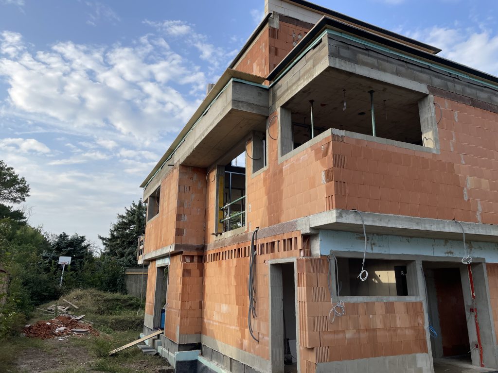 Das Bild zeigt den Rohbau eines zweistöckigen Hauses aus roten Ziegeln. Der Bau befindet sich noch in einer frühen Phase, da das Gebäude noch keine Fenster, Türen oder Außenverkleidung hat. Einige Baustellenmaterialien wie Holz und Gerüstteile liegen rund um das Haus verstreut. An der Außenwand sind einige Kabel sichtbar, was darauf hindeutet, dass elektrische Installationen vorbereitet werden. Es ist möglich, dass dieses Gebäude später als Smart Home ausgestattet wird, da solche Installationen oft schon im Rohbau geplant werden.