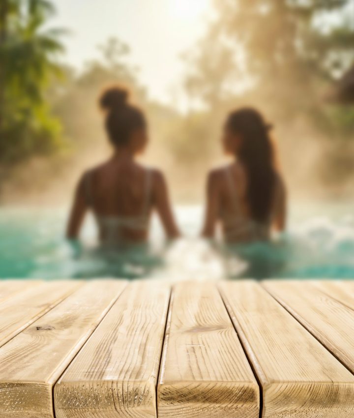 Zwei Frauen sitzen im warmen Wasser eines Pools oder Whirlpools, umgeben von tropischer Vegetation, mit einer hölzernen Poolkante im Vordergrund und Sonnenlicht im Hintergrund.