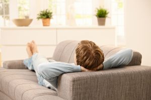 Person liegt entspannt auf einem grauen Sofa, die Hände hinter dem Kopf verschränkt, mit Blick in einen hellen Raum mit Pflanzen und dekorativen Elementen im Hintergrund.