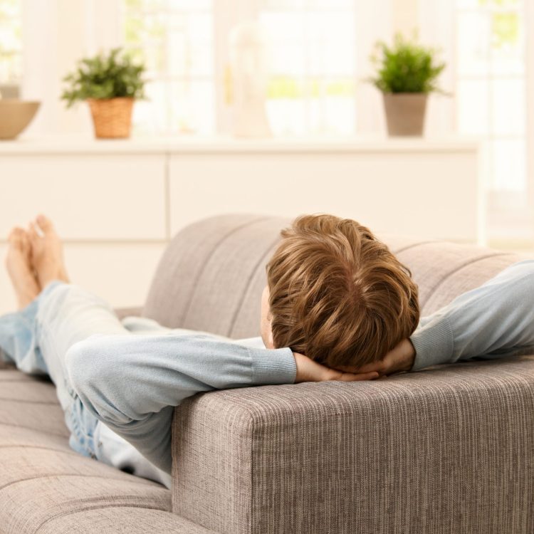 Person liegt entspannt auf einem grauen Sofa, die Hände hinter dem Kopf verschränkt, mit Blick in einen hellen Raum mit Pflanzen und dekorativen Elementen im Hintergrund.