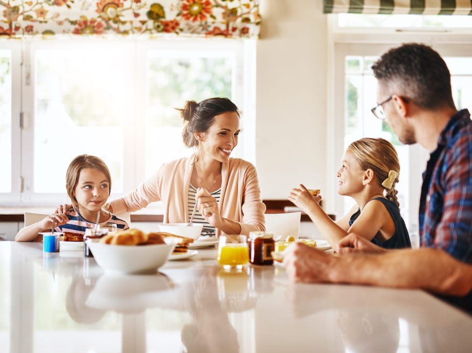 Familie sitzt gemeinsam am Frühstückstisch in einer hellen Küche, isst und lacht, mit verschiedenen Speisen und Getränken wie Croissants, Saft und Marmelade auf dem Tisch.