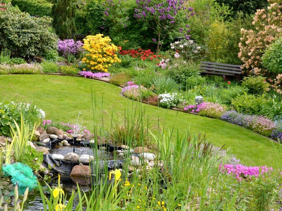 Farbenprächtiger Garten mit gepflegtem Rasen, blühenden Blumenbeeten, kleinen Bäumen, Teich mit Springbrunnen und Sitzbänken zwischen üppiger Vegetation.