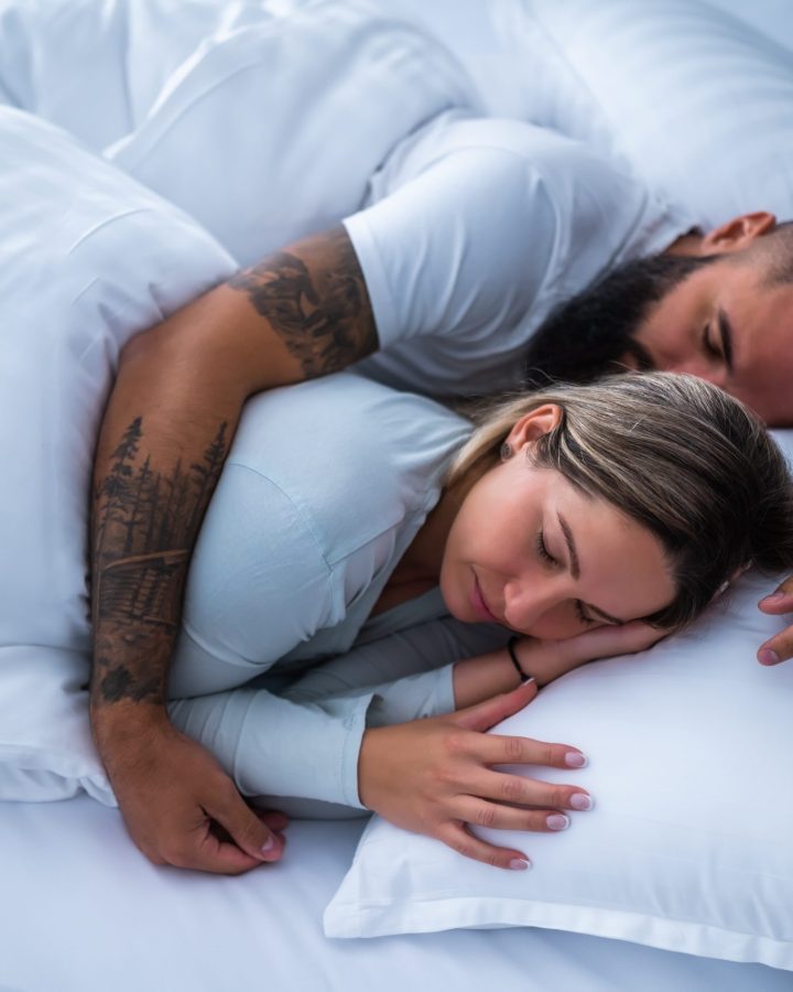 Paar liegt eng umschlungen im Bett, beide schlafen friedlich unter einer weißen Decke auf weißen Kissen.