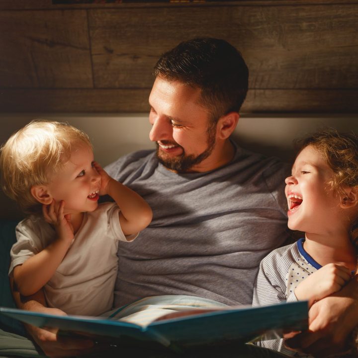 Vater sitzt mit zwei kleinen Kindern im Bett und liest ihnen eine Geschichte vor, während sie zusammen lachen, bei warmem Licht einer Nachttischlampe.