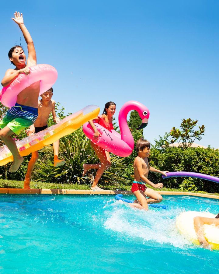 Kinder springen fröhlich mit bunten Schwimmreifen und Poolnudeln in einen Außenpool, während andere im Wasser spielen, umgeben von sonnigem Garten mit blühenden Pflanzen.