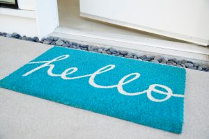 Türkisfarbene Fußmatte mit dem weißen Schriftzug „hello“ liegt vor einer geöffneten Haustür auf einem hellen Boden mit Zierkies am Rand.