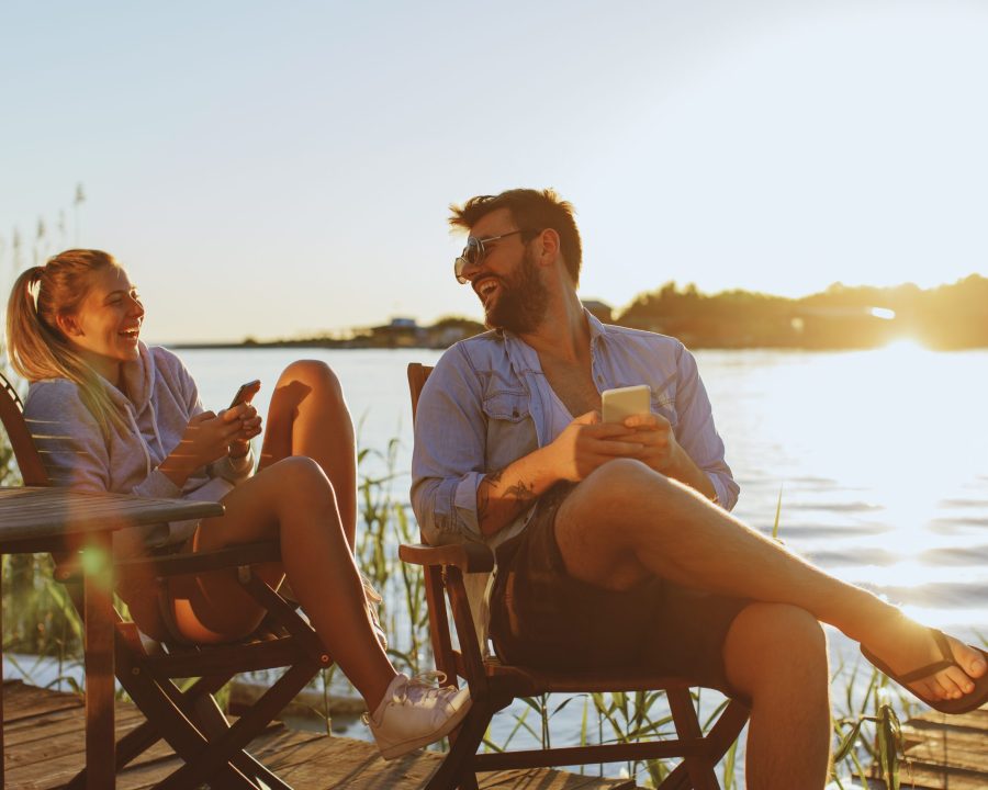 Mann und Frau sitzen entspannt auf Stühlen an einem Steg am Wasser, lachen miteinander und halten Smartphones, während die Sonne im Hintergrund untergeht.