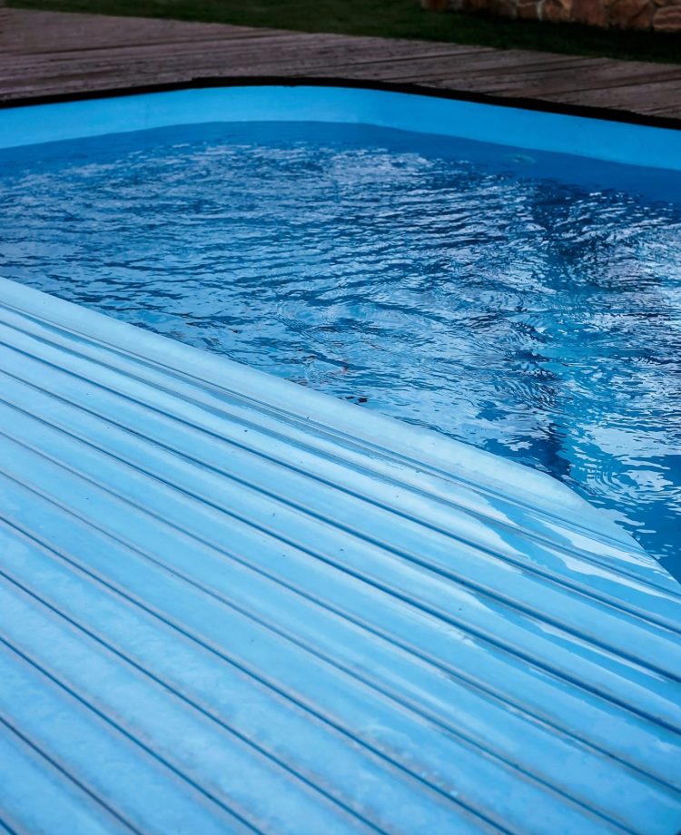 Teilweise geschlossener Pool mit blauer Abdeckung, umgeben von Holzplanken und Steinmauer im Hintergrund.