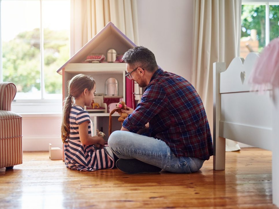 Vater sitzt mit seiner Tochter auf dem Boden im Kinderzimmer und spielt gemeinsam mit einem Puppenhaus und Stofftieren, bei Tageslicht durch große Fenster.