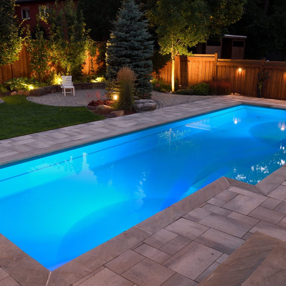 Beleuchteter Swimmingpool im Garten bei Dämmerung, umgeben von Steinplatten, Rasen, Bäumen und einer Holzterrasse mit stimmungsvoller Außenbeleuchtung.