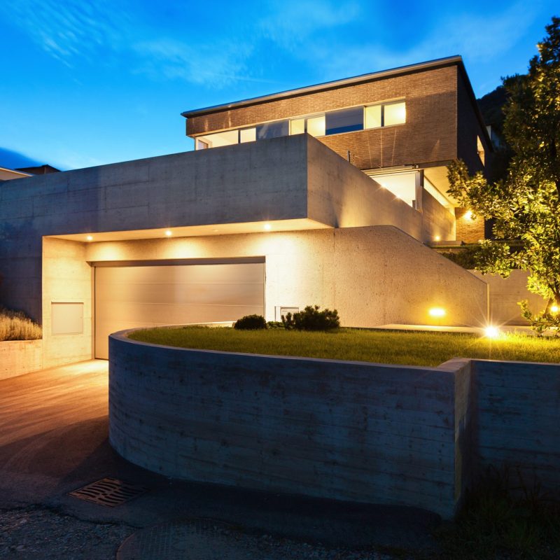 Modernes Wohnhaus mit Beton- und Glasarchitektur, beleuchtet bei Dämmerung, mit Garage, Auffahrt und gepflegtem Vorgarten.